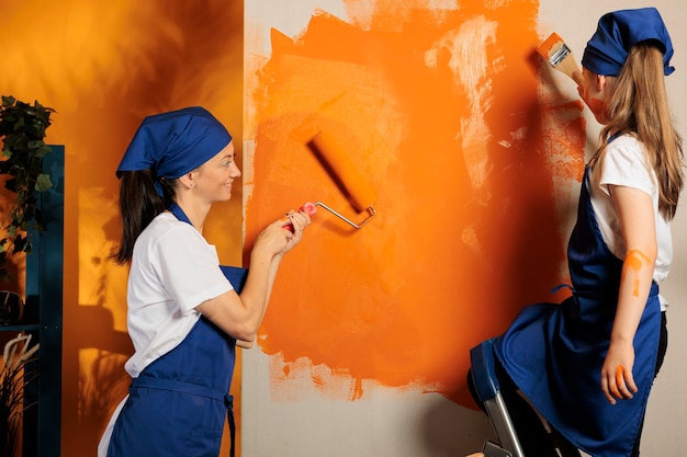 Família caucasiana usando a cor laranja nas paredes, criança na superfície da pintura da escada com pincel e mãe usando a ferramenta de pincel de rolo. Pessoas se divertindo com renovação de trabalhos domésticos, decoração de casa.