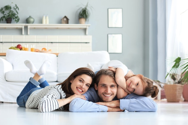 Familia caucásica joven con pose de hija pequeña relajarse en el piso de la sala de estar, sonriente niño niña abrazo abrazar a los padres, mostrar amor y gratitud, descansar juntos en casa.