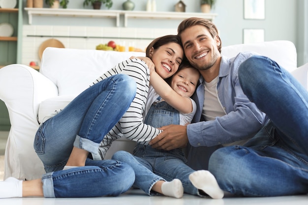 Familia caucásica joven con pose de hija pequeña relajarse en el piso de la sala de estar, sonriente niño niña abrazo abrazar a los padres, mostrar amor y gratitud, descansar juntos en casa.