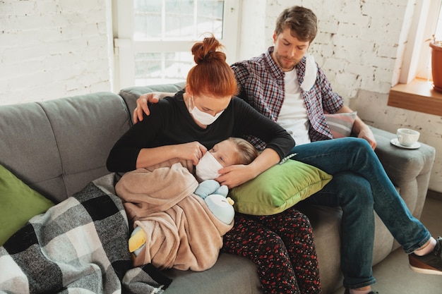 Familia caucásica en mascarillas y guantes aislados en casa con síntomas respiratorios de coronavirus como fiebre, dolor de cabeza, tos en estado leve.