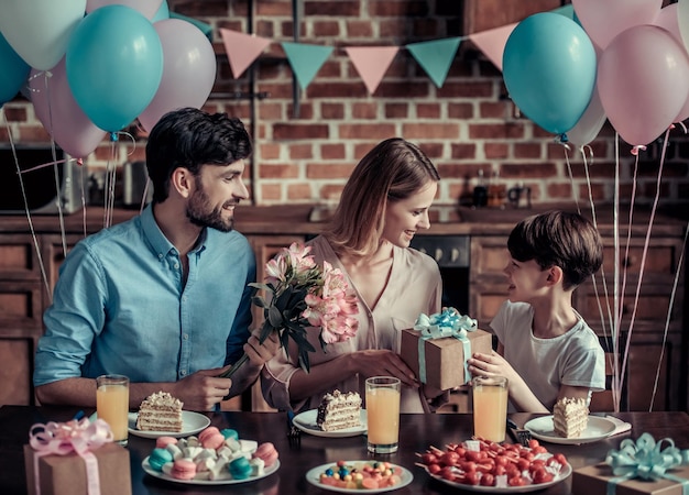 Familia celebrando el cumpleaños de la madre en cocina decorada. Marido con flores, hijo con una caja de regalo, todos están sonriendo