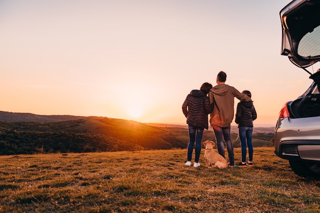 Foto família com cachorro abraçando na colina e olhando o pôr do sol