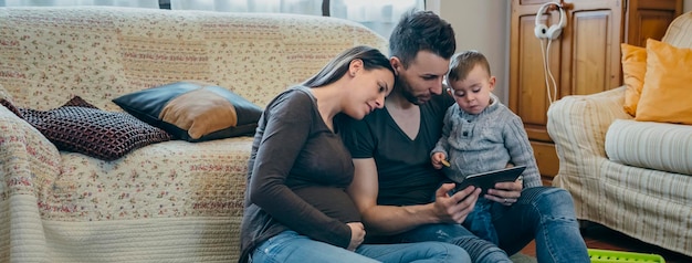 Família com criança pequena e mãe grávida olhando para o tablet sentado no tapete