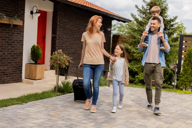 Família com mãe, pai, filho e filha andando com bagagem do lado de fora na varanda da frente de uma casa de tijolos