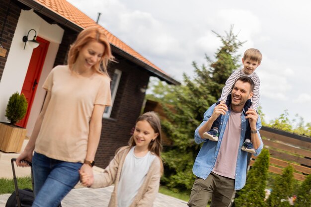 Família com mãe, pai, filho e filha andando com bagagem do lado de fora na varanda da frente de uma casa de tijolos