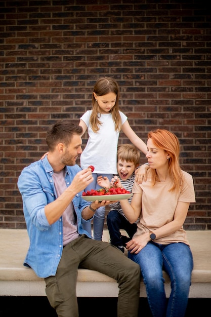 Família com mãe, pai, filho e filha sentados do lado de fora nos degraus da varanda de uma casa de tijolos e comendo morangos