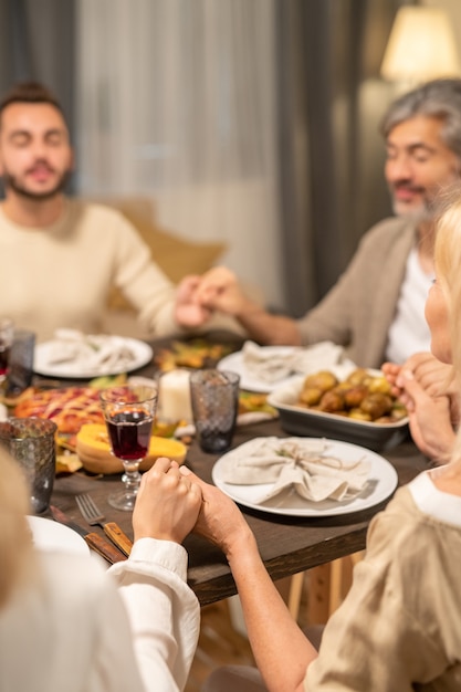 Foto familia contemporánea de cuatro sentados junto a una mesa festiva servida en la sala de estar, sosteniendo de la mano y diciendo gracias al señor mientras mantiene los ojos cerrados