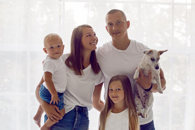 Foto familia de cuatro y un perro de pie junto a una gran ventana en una habitación blanca