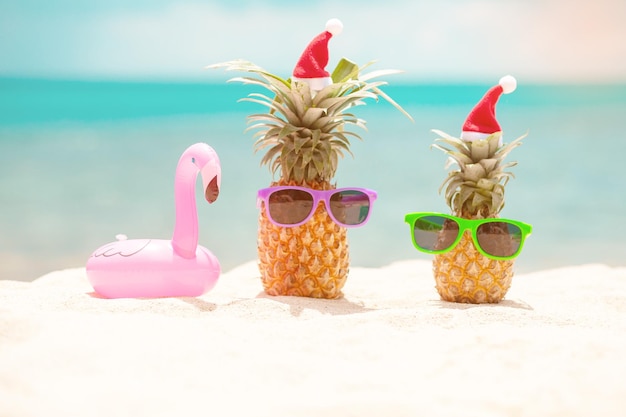 Família de engraçados abacaxis atraentes em elegantes óculos de sol na areia contra o mar azul-turquesa. usando chapéus de natal. conceito de férias de natal e ano novo na praia tropical. feriado familiar. brilhante
