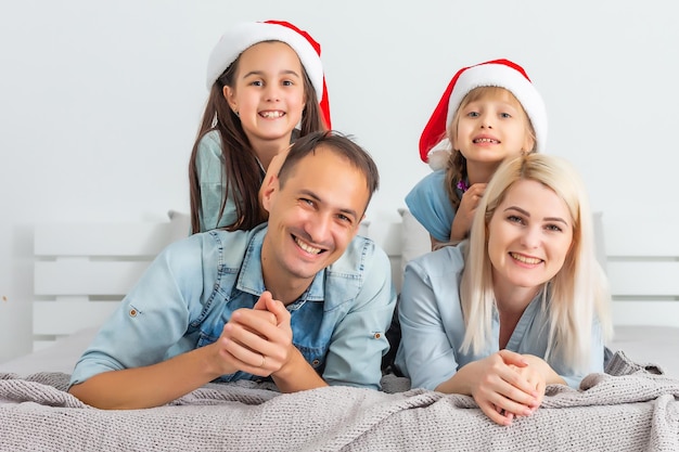 Família de Natal deitada na cama branca com chapéus vermelhos. Pais e filhos felizes