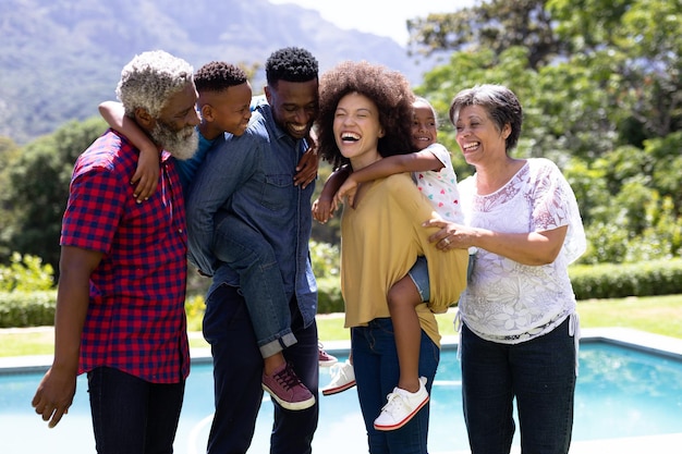 Família de raça mista de várias gerações aproveitando seu tempo em um jardim com piscina, de pé à beira da piscina, abraçando-se, olhando um para o outro e sorrindo, os pais estão segurando seus filhos nas costas