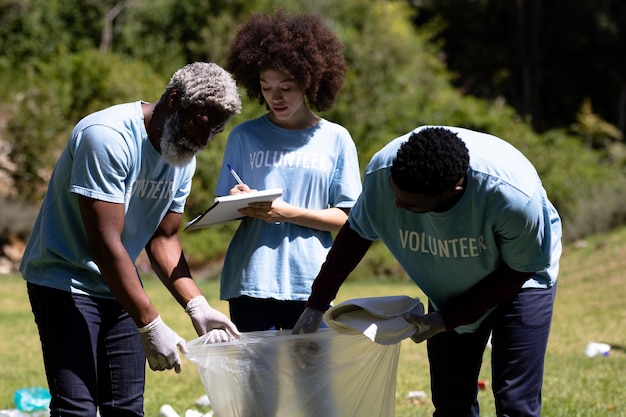 Família de raça mista de várias gerações aproveitando seu tempo em um jardim, todos vestindo camisetas azuis voluntárias, coletando lixo, em um dia ensolarado