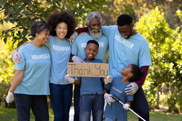 Família de raça mista de várias gerações passando tempo juntos, todos vestindo camisetas azuis de voluntários e luvas de proteção, coletando lixo, abraçando-se, segurando uma placa de voluntário