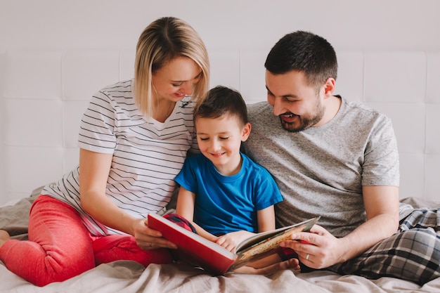 Família de três pessoas sentada na cama no quarto lendo um livro mãe pai menino filho passando tempo juntos