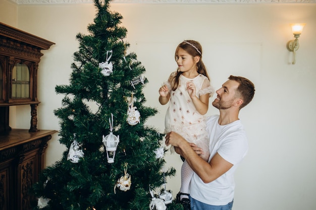 Familia decorando un árbol de Navidad Hombre joven con su hija sobre sus hombros ayudándola a decorar el árbol de Navidad