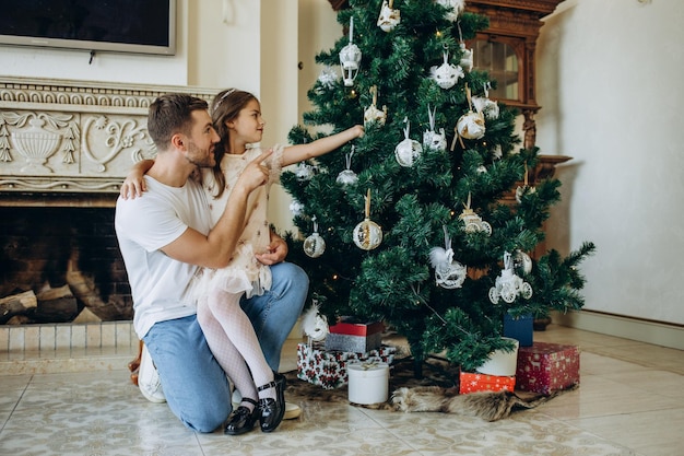 Familia decorando un árbol de Navidad Hombre joven con su hija sobre sus hombros ayudándola a decorar el árbol de Navidad