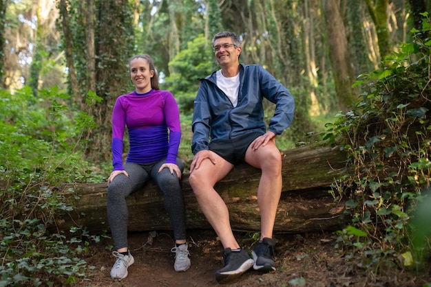 Familia deportiva feliz descansando después de entrenar en el bosque. hombre y mujer caucásicos sentados en un registro sonriendo. deporte, naturaleza, concepto de hobby.