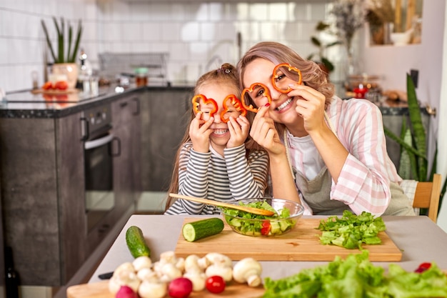La familia se divierte mientras se cocina en la cocina, adorable mujer con niña cortando verduras frescas, sonríe, disfruta del proceso