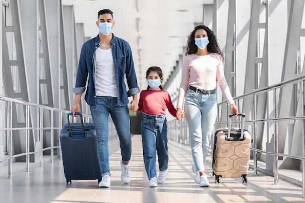 Família do Oriente Médio de três usando máscaras médicas andando com bagagem no aeroporto