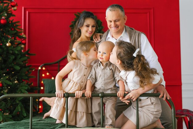 Família elegante com três filhos está sentada em uma cama perto de uma árvore de Natal Um feriado de Natal aconchegante As irmãs beijam seus irmãos, os pais olham para eles e sorriem