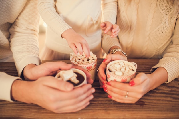 Família em suéteres brancos cozinha e bebe chocolate com marshmallows Closeup mãos e xícaras