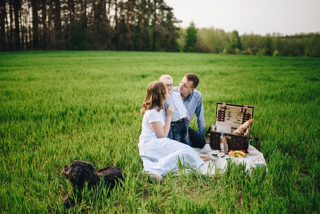 Família em um piquenique na floresta. No campo. Cesta de piquenique com comida. O cachorro corre e quer brincar. Uma criança de óculos. Cor azul na roupa. Para comer ao ar livre. Tempo com a família.