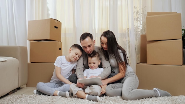 Familia emocionada se sienta entre cajas en el piso y mira la tableta