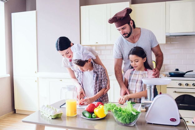 La familia está de pie en la cocina y cocinando. Guy ayuda a la niña a cortar el pepino.