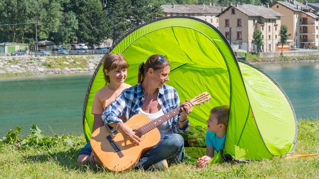 Una familia feliz acampando en el río, tocando la guitarra y cantando juntos una canción en la carpa.