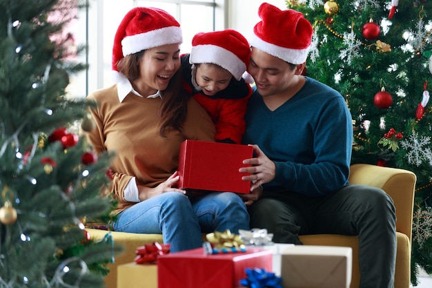 Família feliz asiática e menina abrem a caixa de presente mágica com árvore de Natal na sala decorada para o festival de Natal. Gorros de Papai Noel sentado no sofá. Caixa de presente de Natal do conceito.