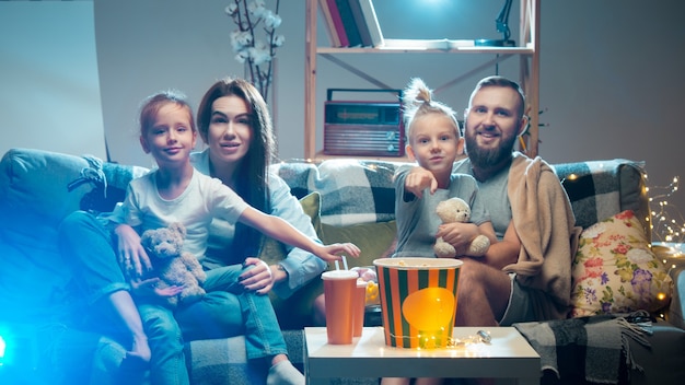 Família feliz assistindo a filmes no projetor de tv com pipoca à noite em casa, mãe, pai e filhos