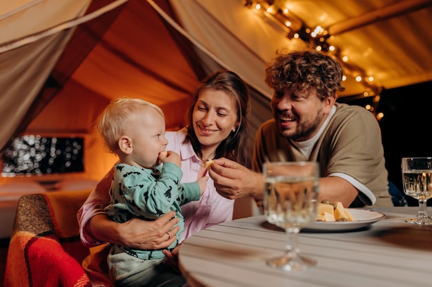 Familia feliz con un bebé encantador cenando y pasando tiempo juntos en glamping en la noche de verano cerca de una acogedora hoguera Tienda de campaña de lujo para recreación al aire libre y recreación Concepto de estilo de vida