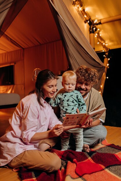 Familia feliz con un bebé encantador jugando y leyendo un libro juntos en glamping en la noche de verano cerca de una acogedora hoguera Tienda de campaña de lujo para recreación al aire libre y recreación Concepto de estilo de vida