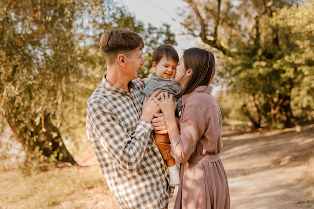 Familia feliz caminando por la carretera en el parque. Padre, madre sosteniendo a su hijo en las manos y juntos. Vista trasera. Concepto de lazos familiares.