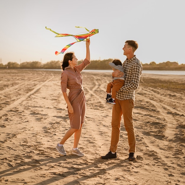 Familia feliz caminando en la playa del río. Padre, madre sosteniendo a su hijo en las manos y jugando con la cometa.