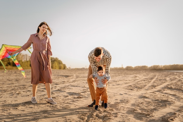 Familia feliz caminando en la playa del río. Padre, madre sosteniendo a su hijo en las manos y jugando con la cometa.