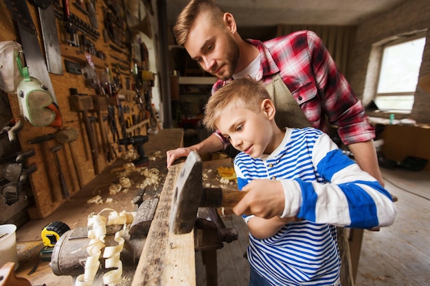 familia feliz, carpintería, carpintería y concepto de personas - padre e hijo pequeño con martillo clavando clavos en tablones de madera en el taller