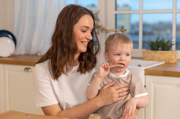 Familia feliz en casa madre alimentando al bebé en la cocina niño pequeño con cara graciosa desordenada come sano fo