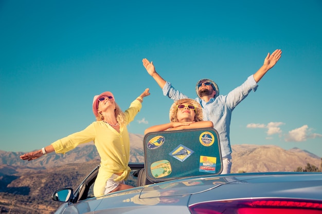 Familia feliz con coloridos sombreros y maleta en el techo de su auto convertible con fondo de montaña