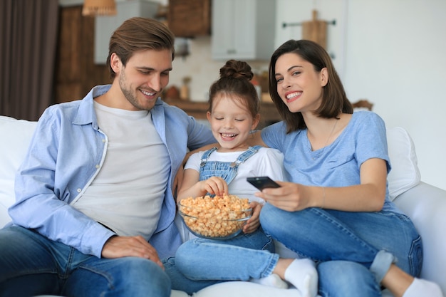 Família feliz com a criança sentada no sofá assistindo tv e comendo pipoca, jovens pais abraçando a filha relaxando no sofá juntos.