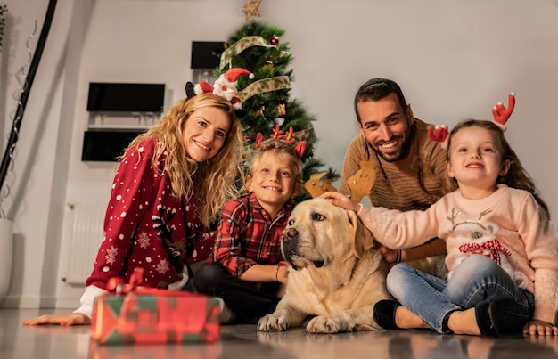 Família feliz com seu cachorro labrador sentado no chão comemorando o natal olhando para a câmera