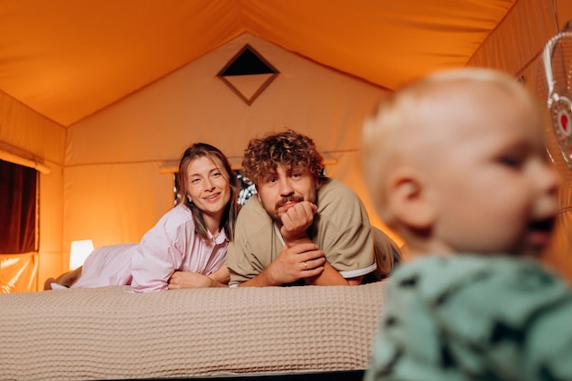 Família feliz com um bebê adorável brincando e passando um tempo juntos em um glamping aconchegante na noite de verão, deitado na cama Barraca de acampamento de luxo para recreação e recreação ao ar livre