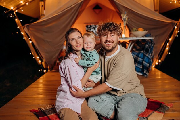 Família feliz com um bebê adorável relaxando e passando um tempo juntos em glamping na noite de verão perto de uma fogueira aconchegante Barraca de acampamento de luxo para recreação ao ar livre e recreação Conceito de estilo de vida