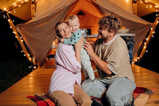 Família feliz com um bebê adorável relaxando e passando um tempo juntos em glamping na noite de verão perto de uma fogueira aconchegante Barraca de acampamento de luxo para recreação ao ar livre e recreação Conceito de estilo de vida