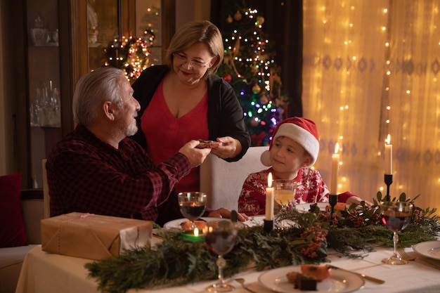 Família feliz comemorando o Natal à noite em casa. Jantar festivo perto da árvore de Natal. É hora de dar presentes. Conceito de amor e união familiar.