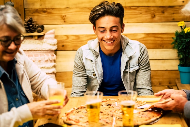 Família feliz de dois idosos e um adolescente em casa ou restaurante comendo pizza e bebendo cerveja juntos - mesa de madeira