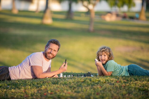 Família feliz de pai homem e filho jogando xadrez na grama verde na paternidade ao ar livre do parque