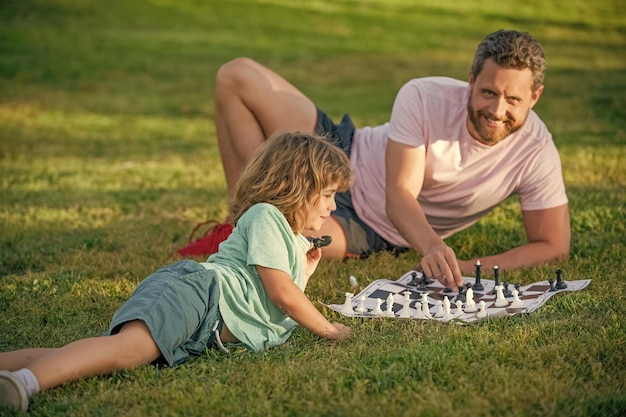 Família feliz de papai e filho jogando xadrez na grama verde no xeque-mate ao ar livre do parque