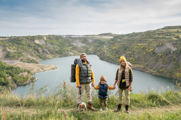 Família feliz de um jovem casal e seu filho pequeno em roupas casuais aconchegantes, em pé na trilha contra o lago e as montanhas durante a viagem