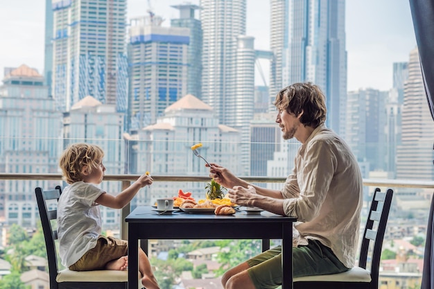 Familia feliz desayunando en el balcón Mesa de desayuno con café, fruta y croissant de pan en un balcón con el telón de fondo de la gran ciudad